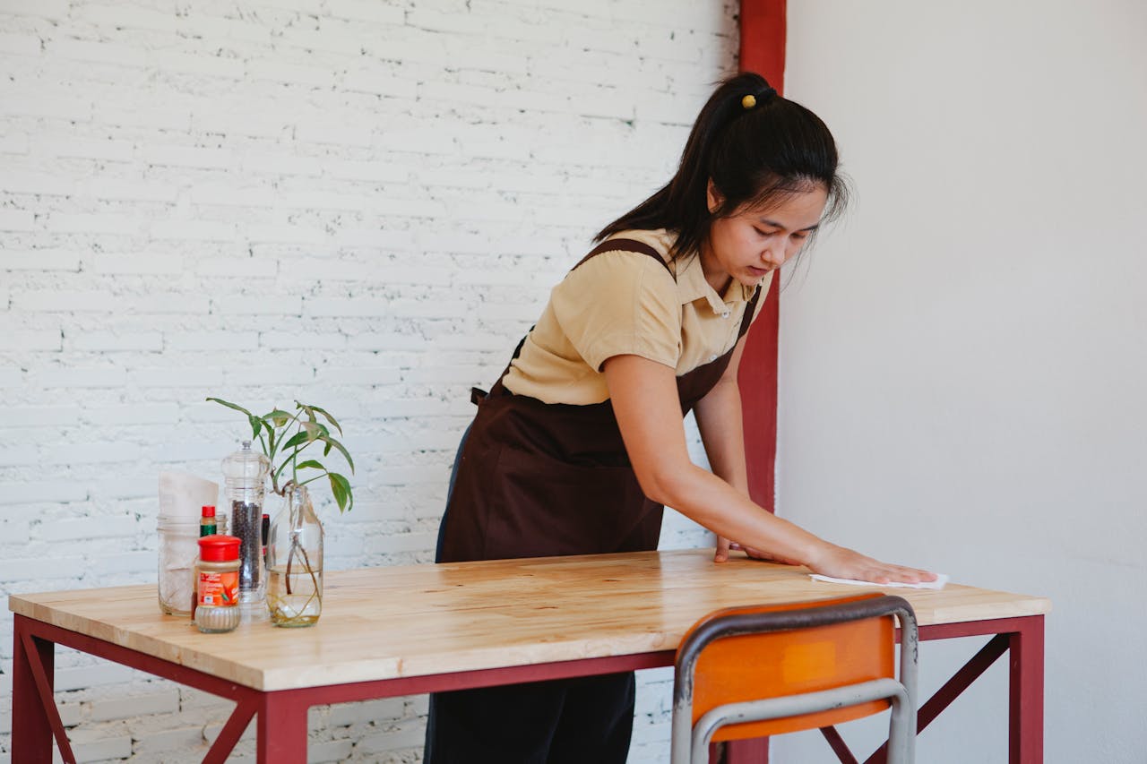 gallery-01 Asian woman wiping a wooden table in an indoor setting with cleaning items.